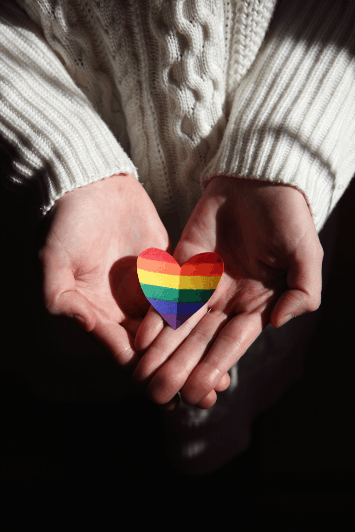 A person wearing a white sweater with outstretched palms, cupping a paper cutout of a heart shape. The heart has rainbow stripes.