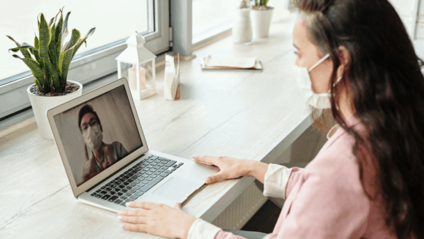 A woman wearing a mask and looking at a mirror reflection of herself on the laptop.