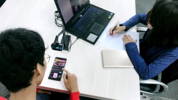 A usability test in progress. One person is viewing an app on a mobile phone, and another person is observing the video feed of the test on a laptop screen, taking notes.