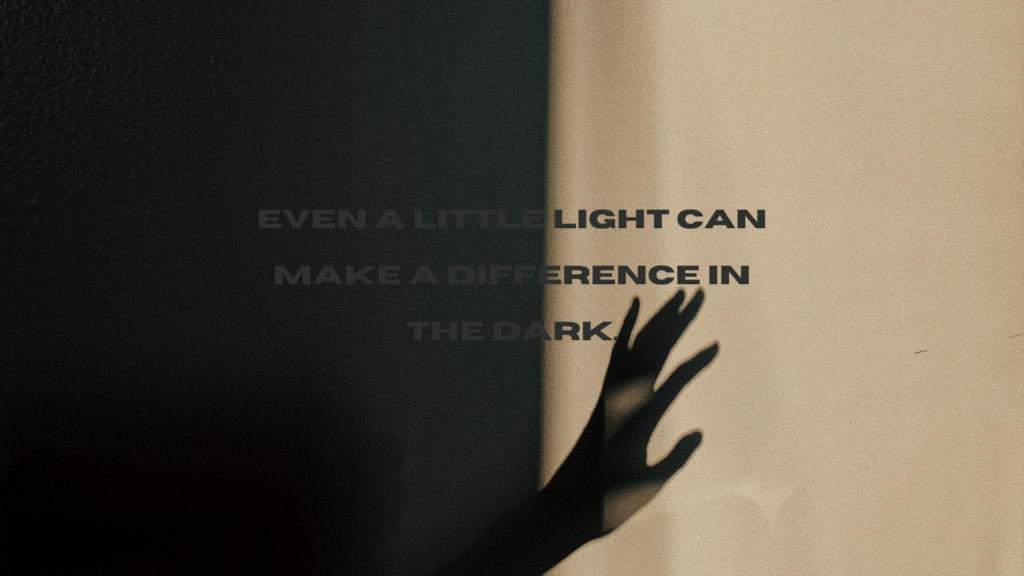 A person reaching out in a room with poor light. The caption on the image reads, "Even a little light can make a difference in the dark." The caption is written in dark colours, making it very difficult to read against the dark background.